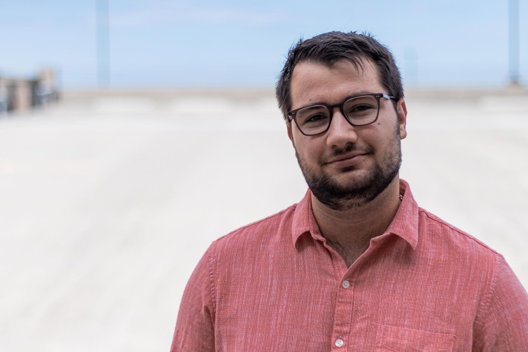 Man with beard and glasses stands in dester with pink collard shirt.