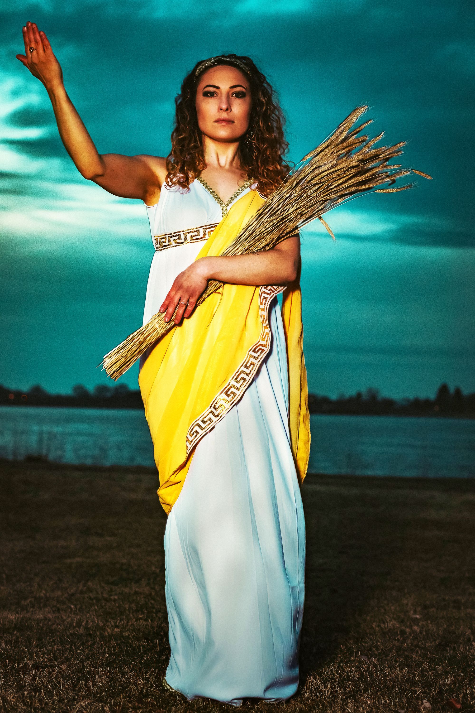 Photograph of woman in toga holding wheat in one arm and holding up her other arm, in front of a river