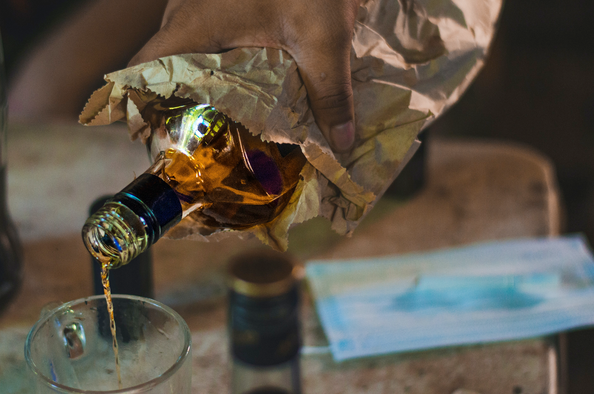 Photo of a hand pouring a bottle of alcohol into a glass. The bottle is inside a paper bag and there is a medical mask in the background