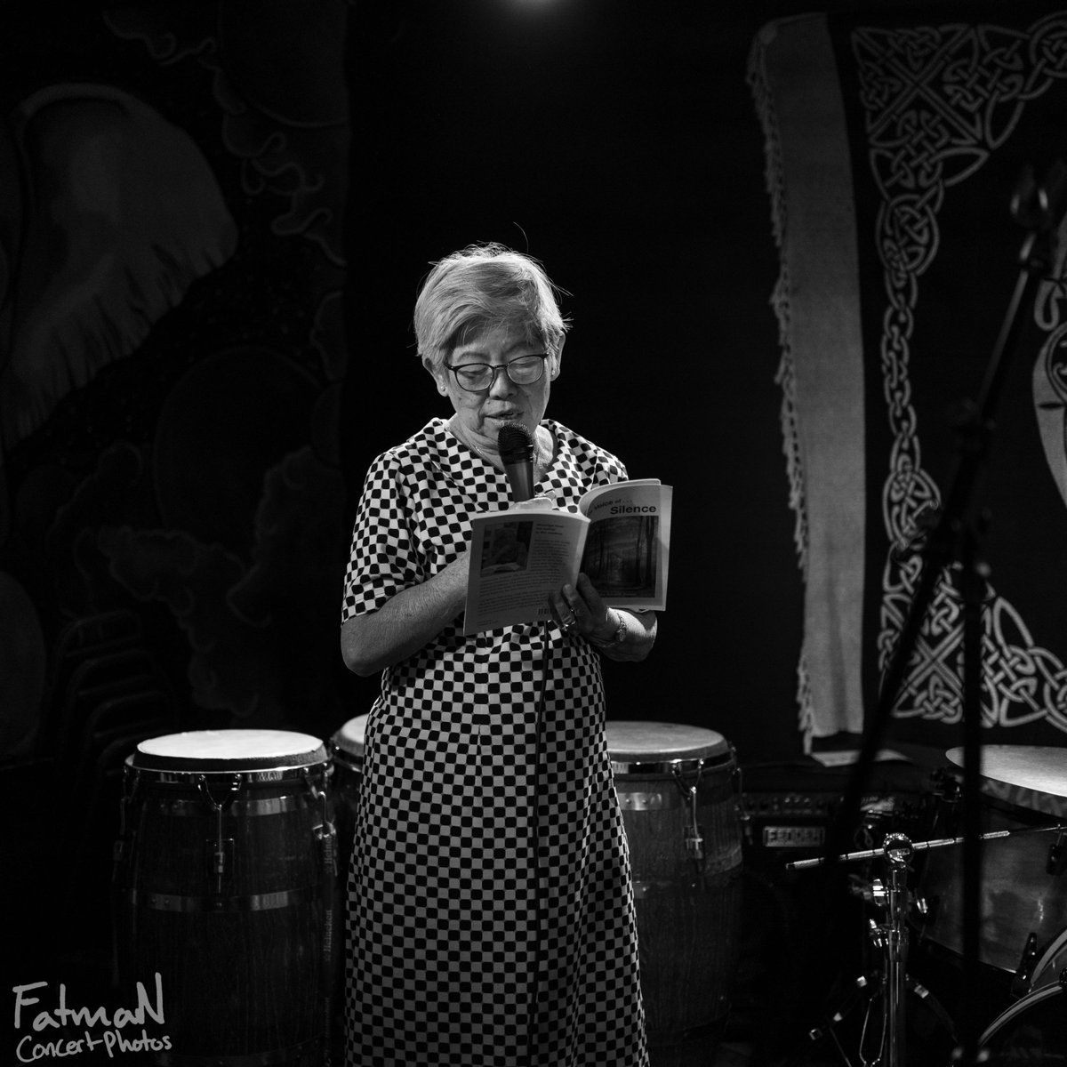 Black and white photo of an older woman with glasses and checkered dress standing on stage reading