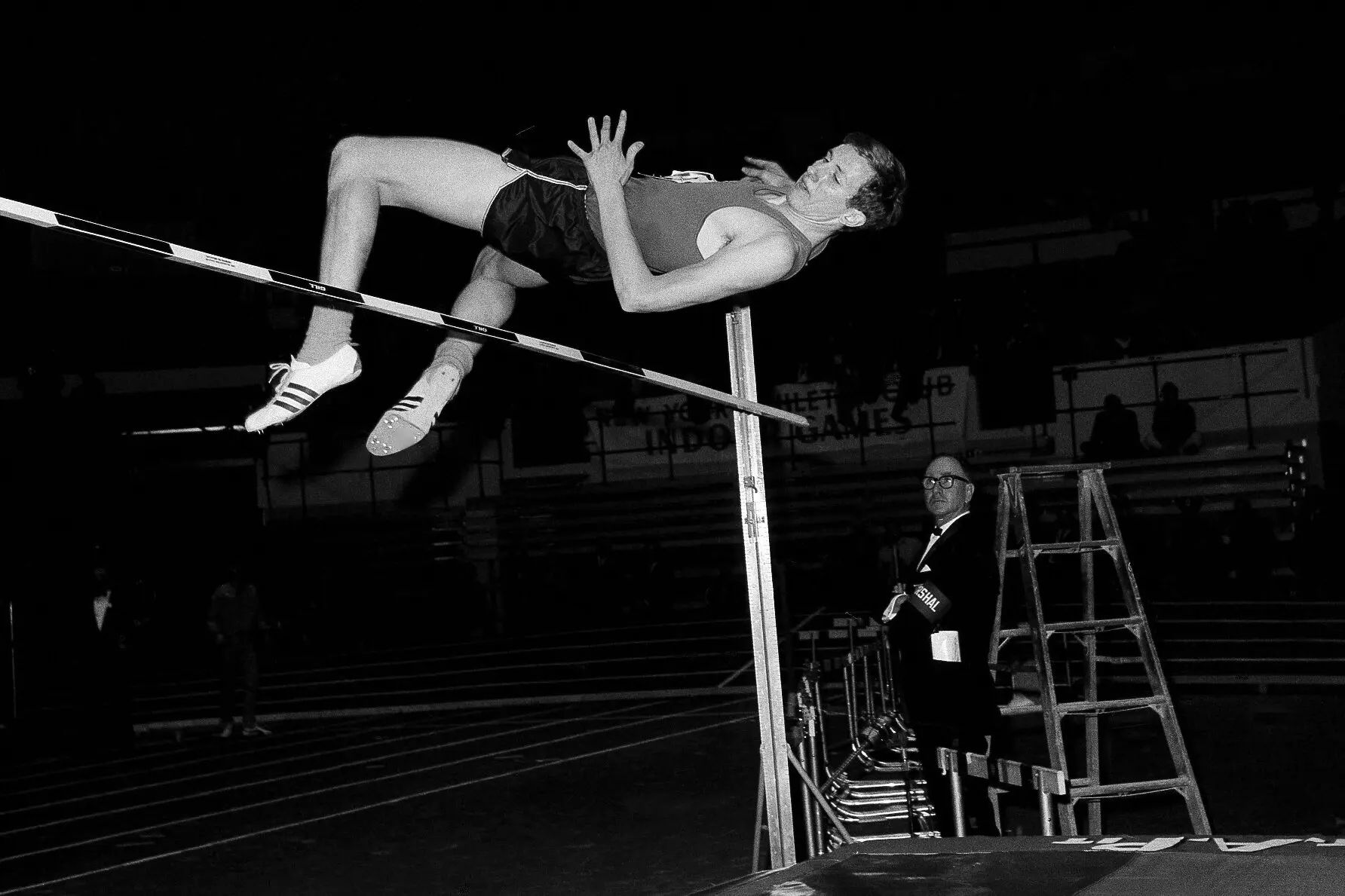 An image of a White man with short hair jumping over a bar backwards, with another man in glasses standing behind watching.
