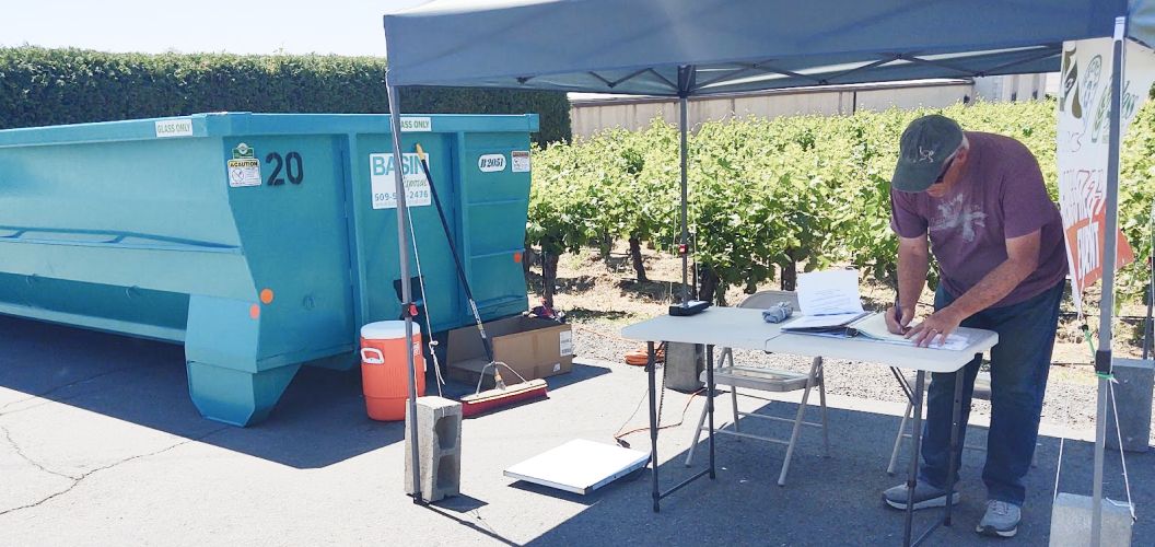 An image of a man under a canopy tent writing something down, it is sunny and there are bushes behind him.