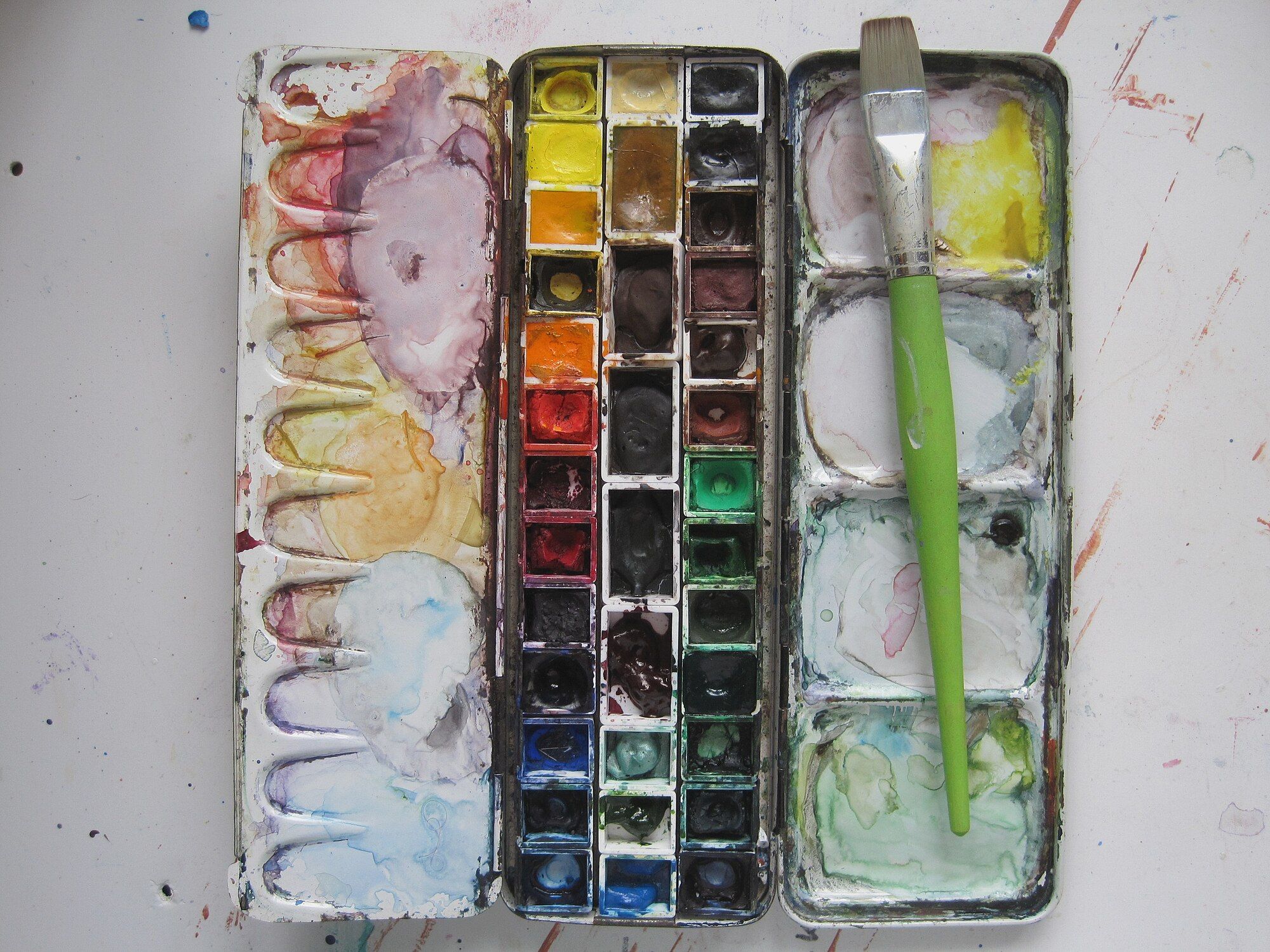 An image of a used watercolor paint tray with a paintbrush laying over it.